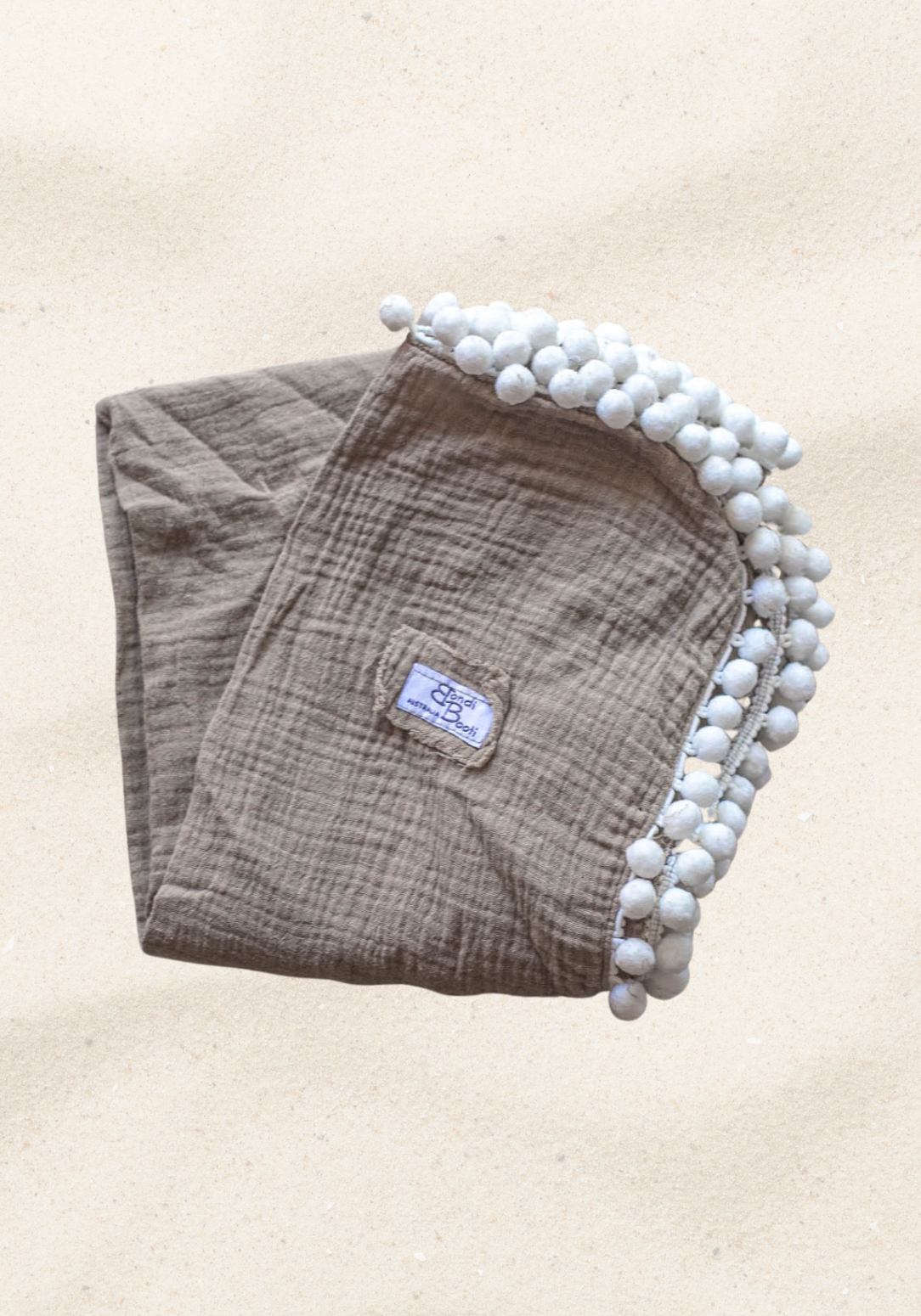 Taupe organic cotton baby blanket with white pom-pom trim, neatly folded with visible Bondi Booti label, displayed on a neutral background.
