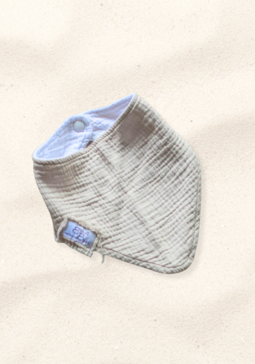 Soft beige organic cotton bandana dribble bib for babies with rounded shape and snap closure, displayed on a sand background