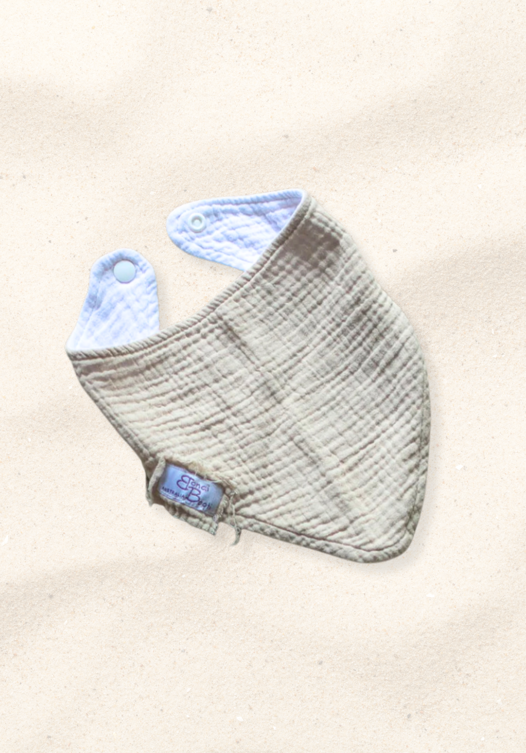 Soft beige organic cotton bandana dribble bib for babies with rounded shape and snap closure, displayed on a sand background