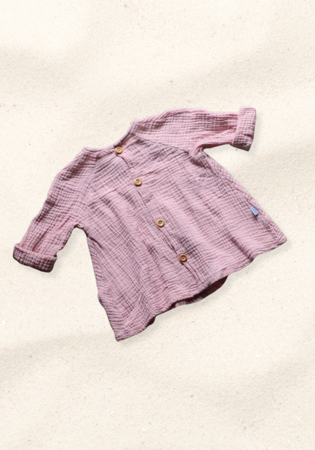 Front view of dusty pink boho muslin baby dress with buttons and folded sleeves on sand background