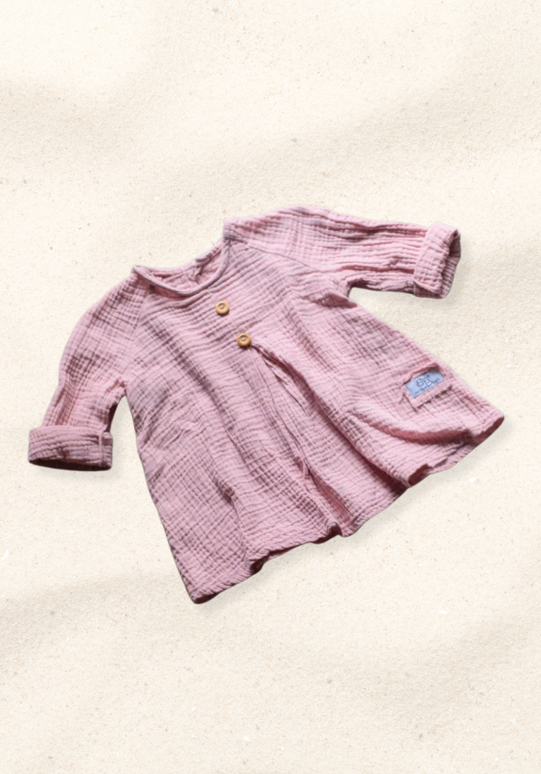 Front view of dusty pink boho muslin baby dress with buttons and folded sleeves on sand background