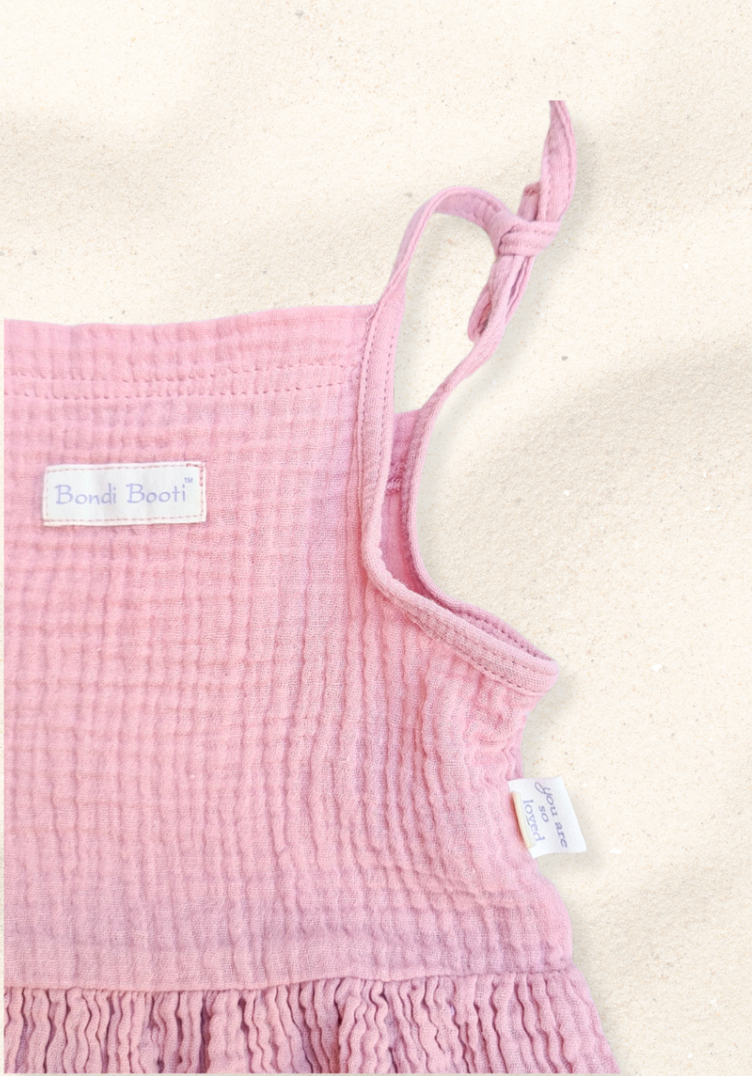 Dusty pink baby picnic dress with ruffled sleeves and gathered waist, made from soft cotton and laid flat on a sand-colored background.