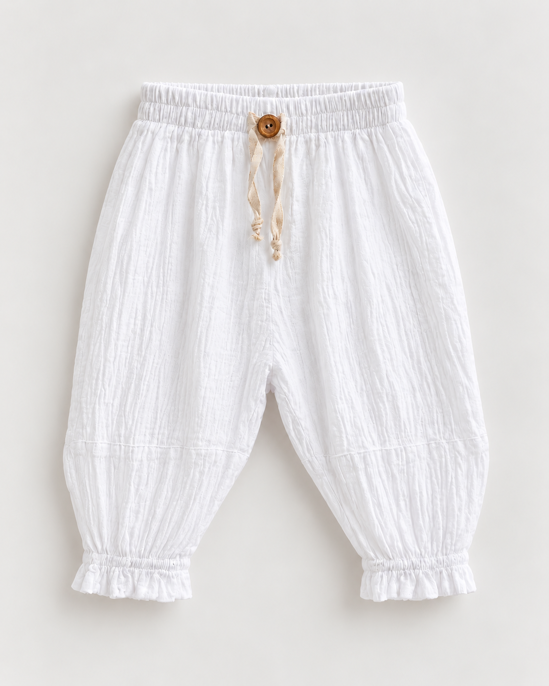 White crinkled cotton harem pants for toddlers, featuring an elastic waistband, a decorative wooden button, a cream drawstring, and ruffled cuffs at the ankles.