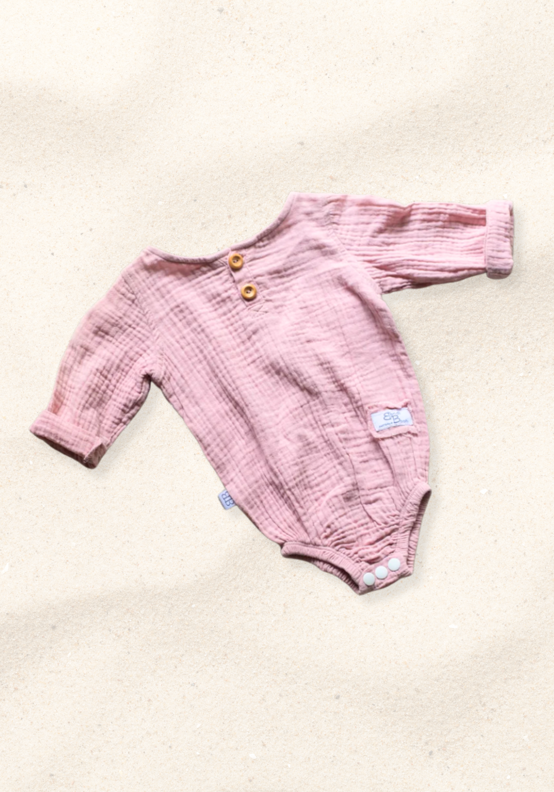 Front view of pink long sleeve baby romper with wooden buttons and soft muslin fabric on sand background