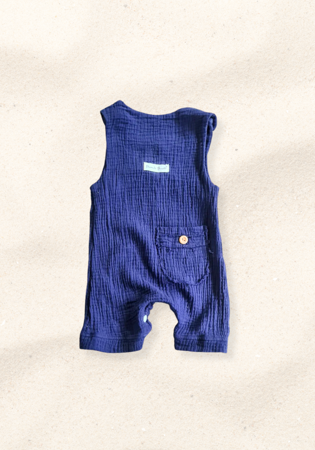 Navy organic cotton baby overall onesie complete with crotch snaps and wooden buttons, displayed on a sand background