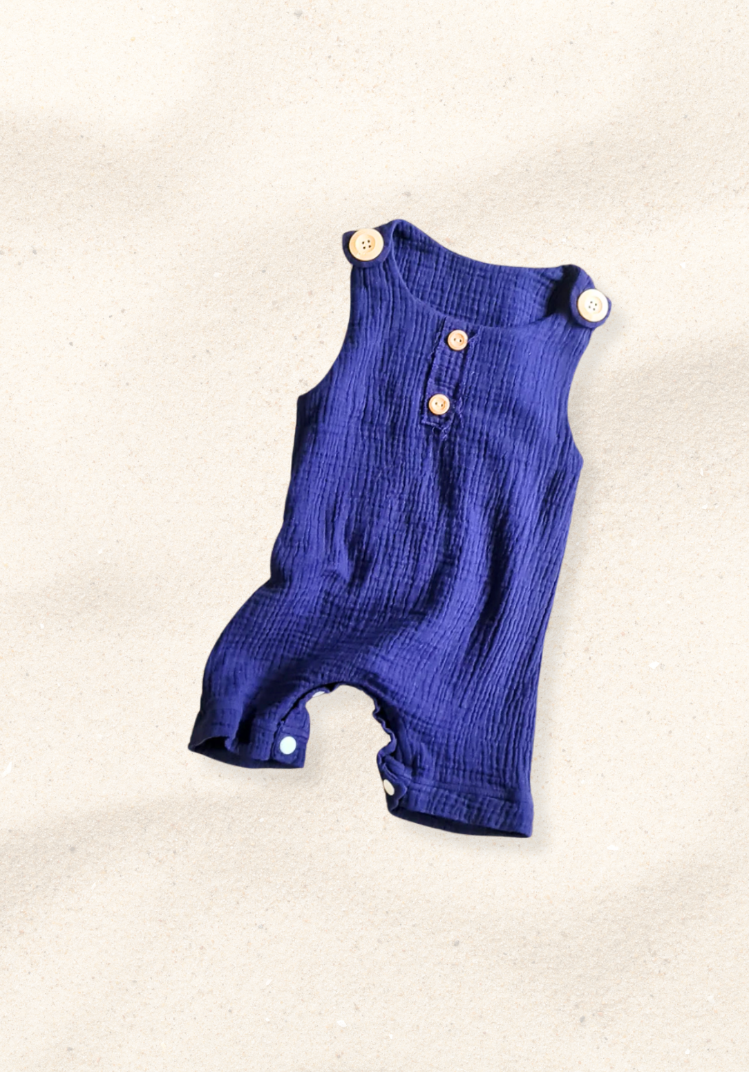 Navy organic cotton baby overall onesie complete with crotch snaps and wooden buttons, displayed on a sand background